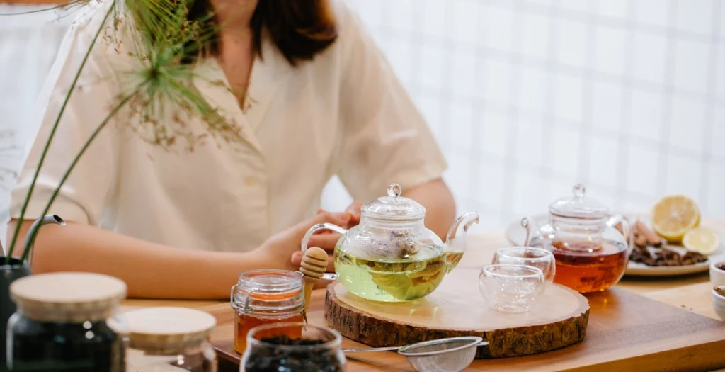 A woman sitting at a wooden table with glass teapots of herbal detox teas and jars of loose ingredients.