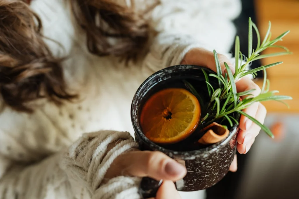 A cozy setting with a cup of detox tea, garnished with lemon, rosemary, and cinnamon held by a woman in a knitted sweater.