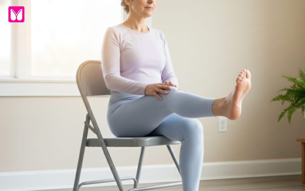 A woman in light purple and blue activewear sits tall on a folding chair, extending one leg straight out in front of her with her foot flexed toward the ceiling. She gently supports the other leg with her hands, demonstrating a seated leg strengthening exercise from a Free 28-Day Chair Yoga for Seniors program.